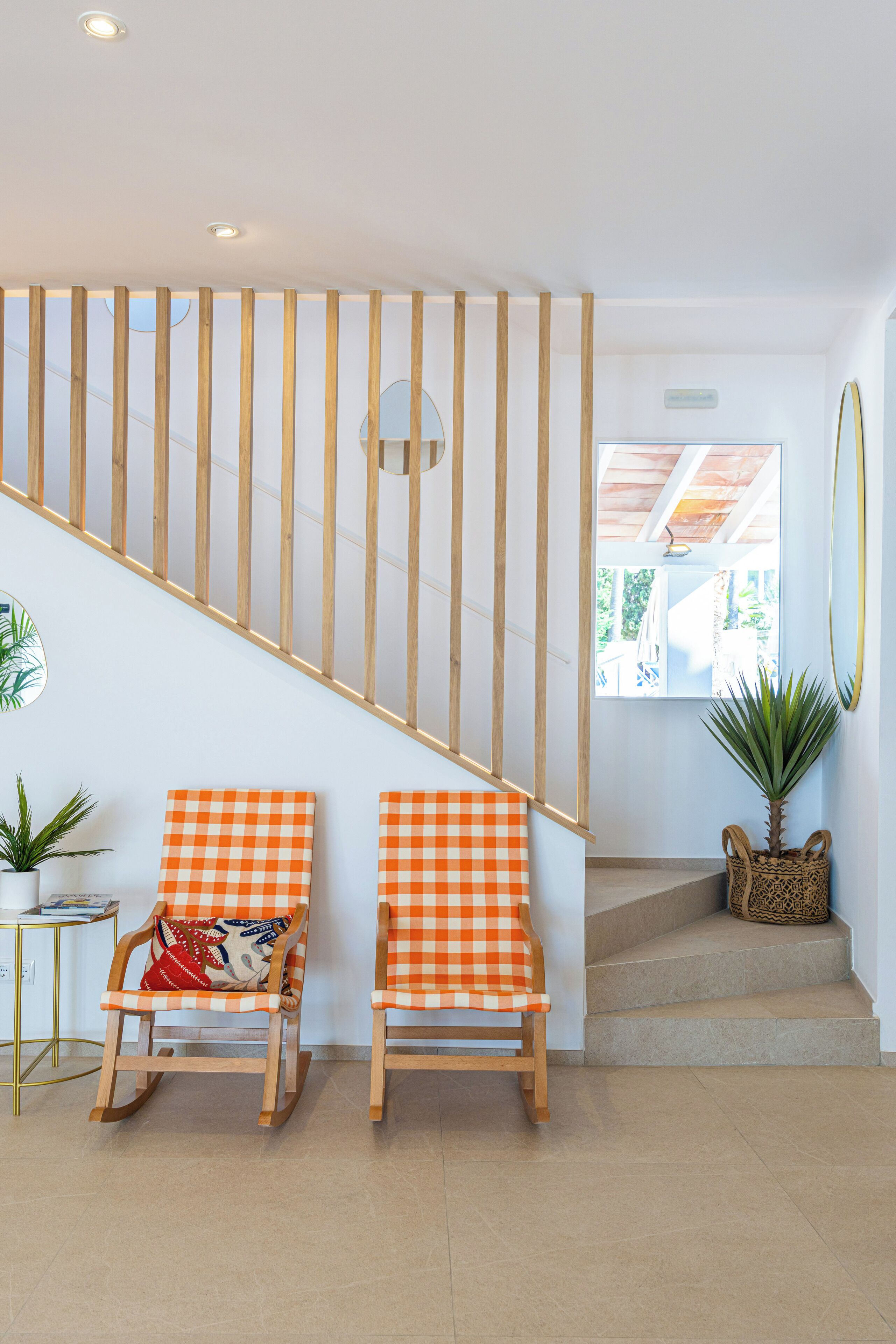 Un escalier avec des rampes en bois, deux fauteuils à bascule recouverts de housses à carreaux orange et une plante en pot sur le palier.