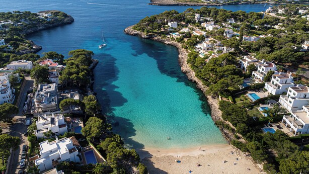 Vue aérienne d’une ville côtière avec une baie turquoise aux eaux limpides, des bâtiments blancs et une plage de sable.