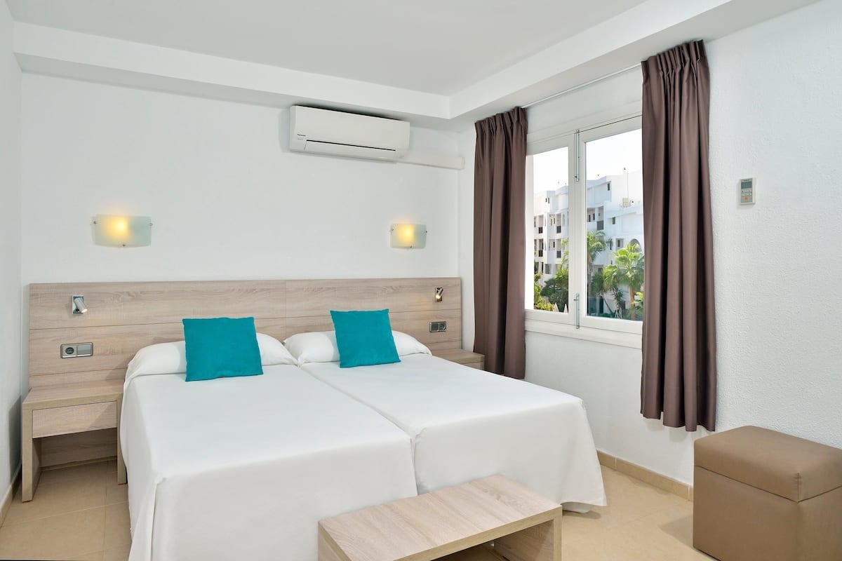 Une chambre d’hôtel avec deux lits, une table de chevet, un petit banc en bois, un appareil de climatisation et une fenêtre avec des rideaux.