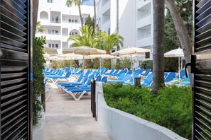 Un área junto a la piscina con sillas reclinables azules y sombrillas, rodeada de vegetación y un edificio con varios balcones.