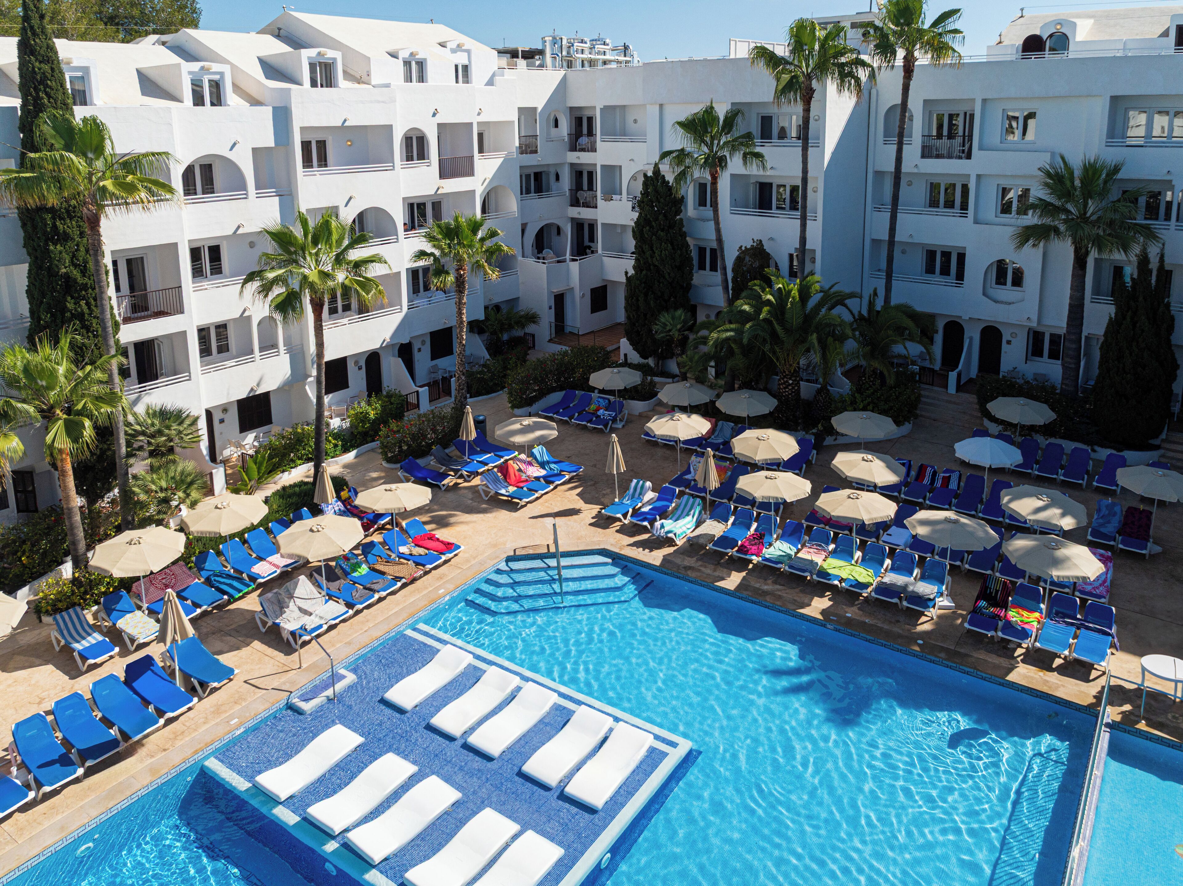 Ein Hotel mit Poolbereich, mehreren Liegestühlen, Sonnenschirmen und Palmen.