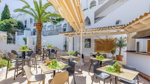 Un espace extérieur aménagé en terrasse, avec des tables et des chaises, entouré de plantes en pot et d’un palmier.