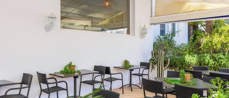 An outdoor seating area with tables and chairs, surrounded by greenery and a large window.