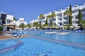 Una piscina con azulejos azules, rodeada de sillas de descanso y sombrillas, con edificios blancos y palmeras al fondo.
