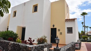 A two-story building with a white facade and a yellow accent wall, featuring a black door and windows, and a stone wall with plants in front.