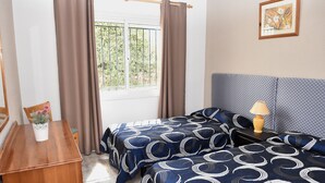 A bedroom with two beds, a wooden dresser, a window with curtains, a nightstand with a lamp, and a framed picture on the wall.