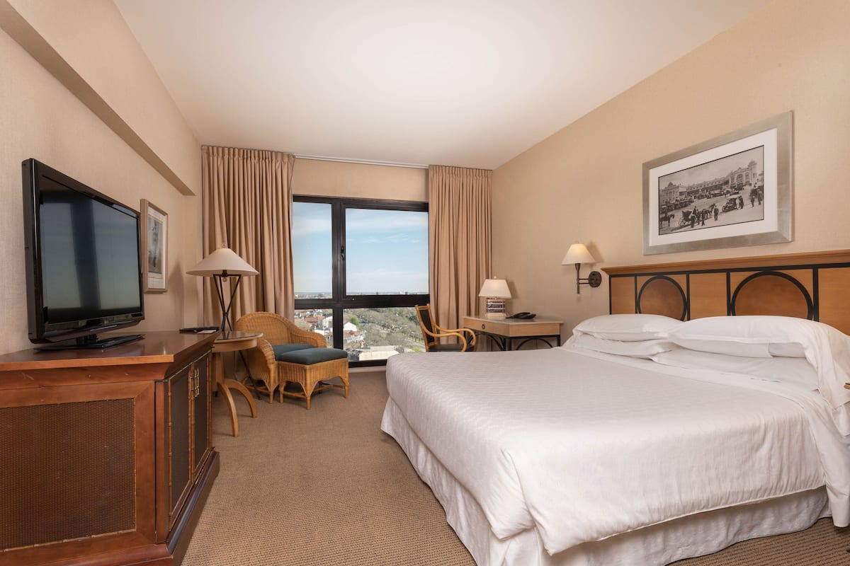 Une chambre d’hôtel comprenant un lit, une télévision, un bureau avec une lampe, une chaise et une fenêtre avec des rideaux.