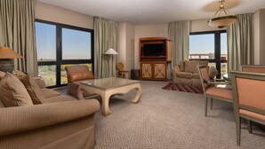 Ein geräumiges Wohnzimmer mit einer Sofagarnitur, einem Couchtisch und einem Fernseher. Große Fenster bieten Blick auf die Stadtlandschaft.