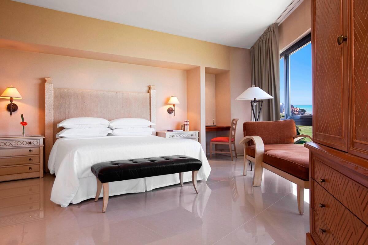 Une chambre d’hôtel avec un grand lit, un banc, une chaise, une table de chevet, une lampe et une vue sur l’extérieur.