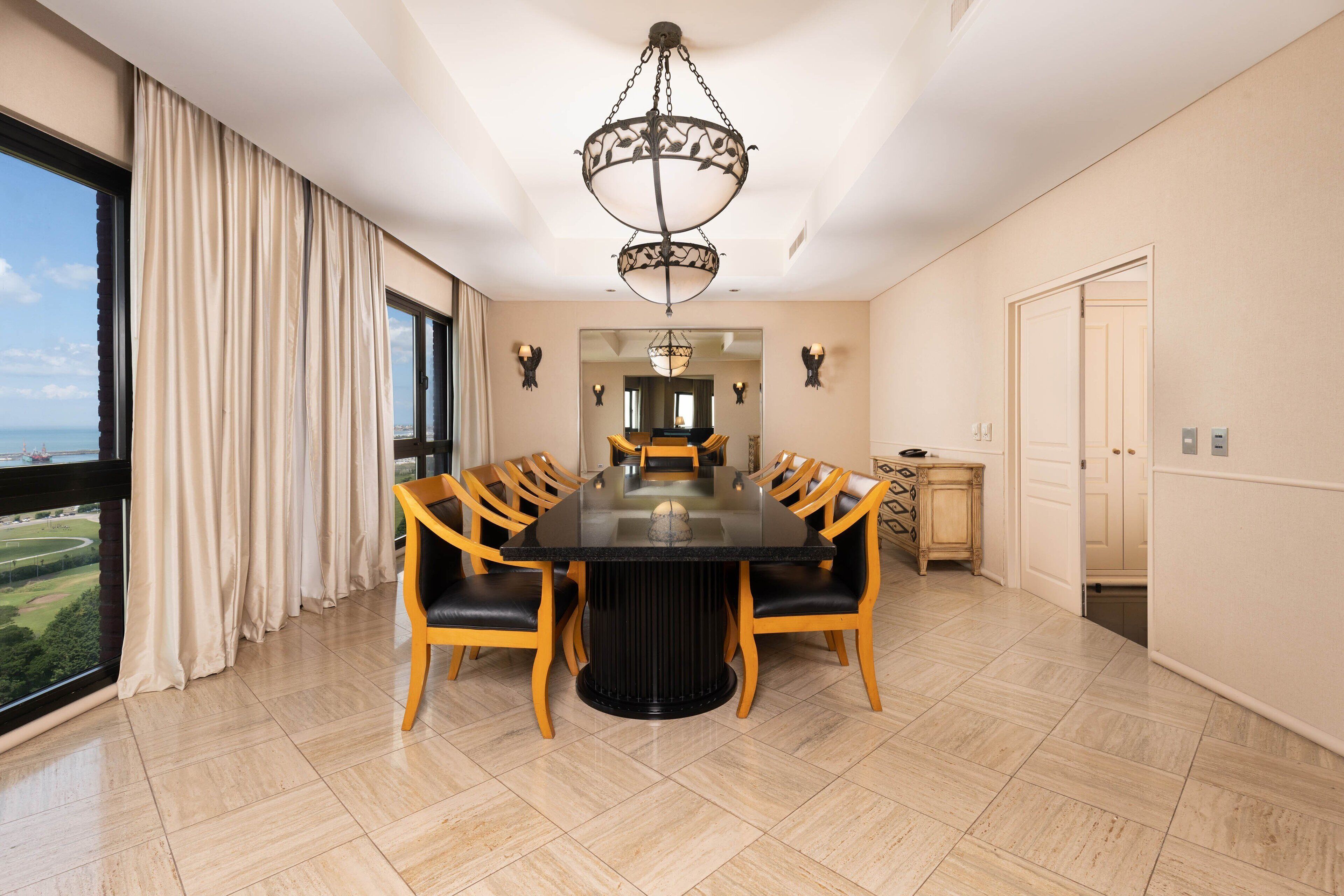 A spacious dining room with a large table, wooden chairs, a chandelier, and a view of the sea.