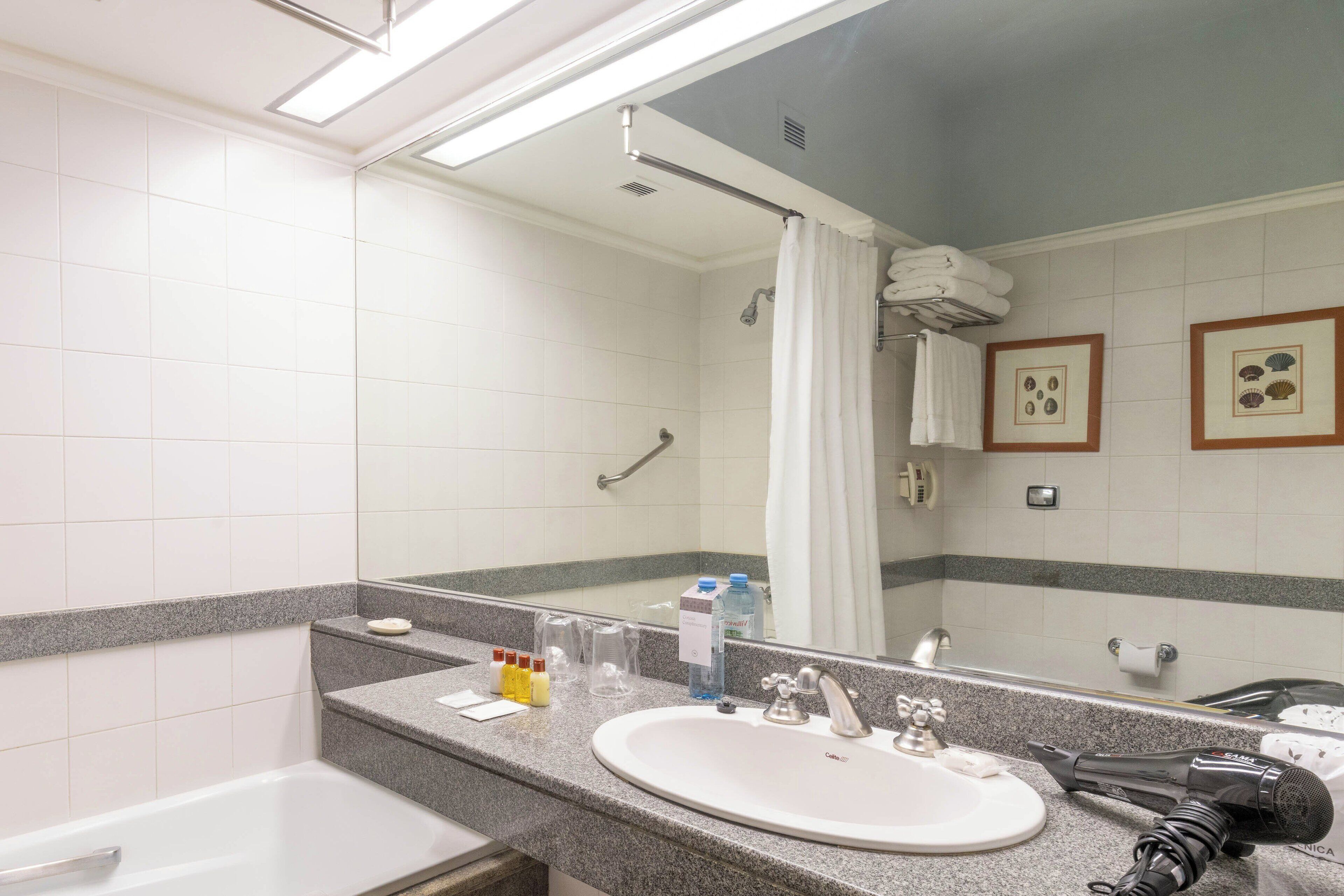 Ein Badezimmer mit Waschbecken, Spiegel und Dusche. Auf dem Waschbecken stehen Pflegeprodukte und ein Föhn.