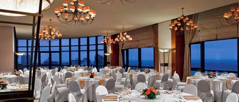 Un salón de banquetes con mesas redondas, manteles blancos y sillas con fundas blancas. Varios candelabros cuelgan del techo.