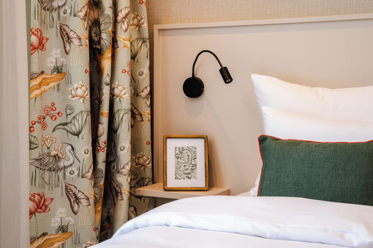 Un letto rifatto con lenzuola bianche, un cuscino verde, un quadro incorniciato su un comodino e una lampada fissata al muro.