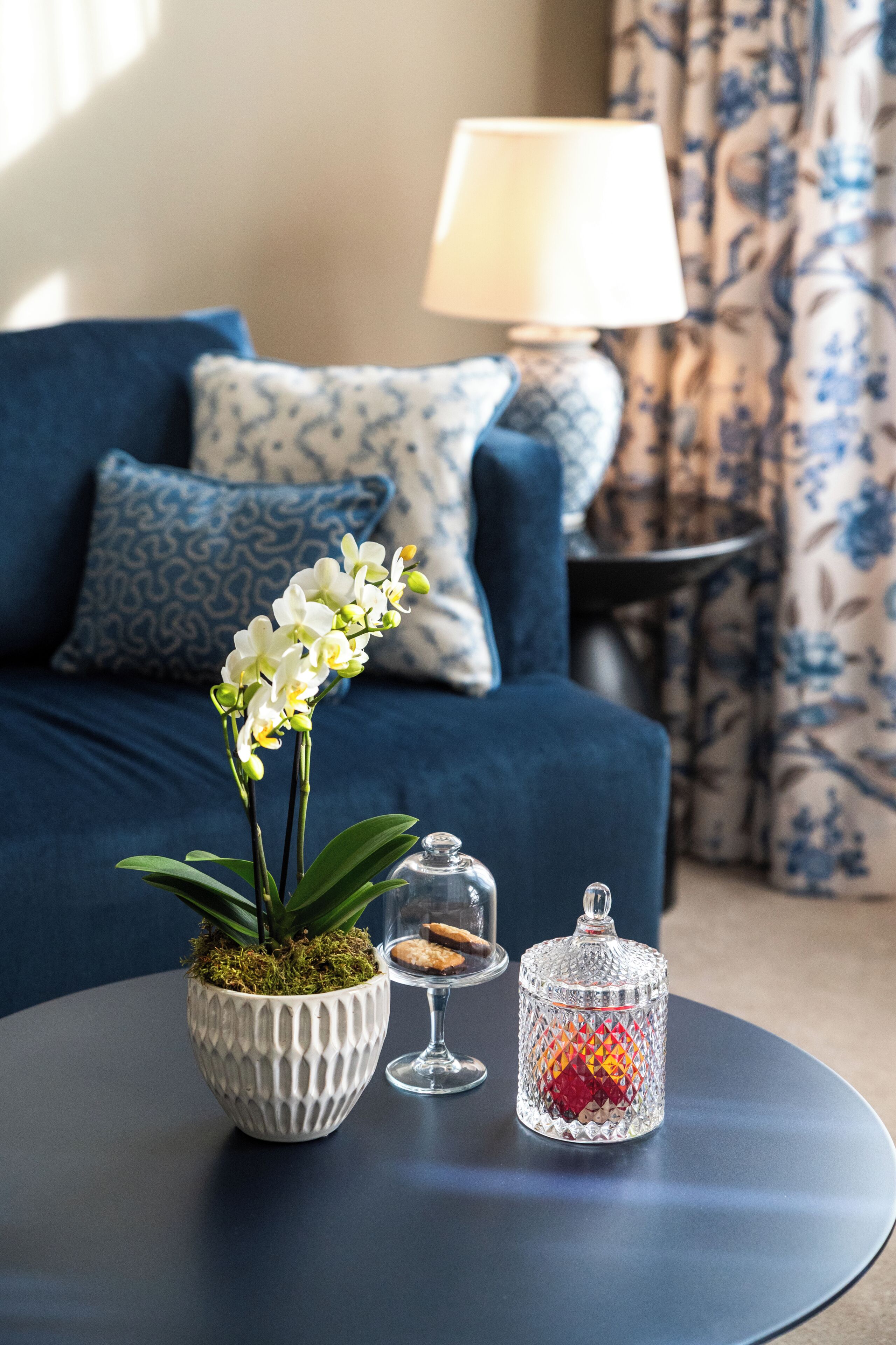 Una stanza con un divano blu, cuscini decorati, un vaso bianco con un'orchidea, un portacandela di vetro e un tavolino.