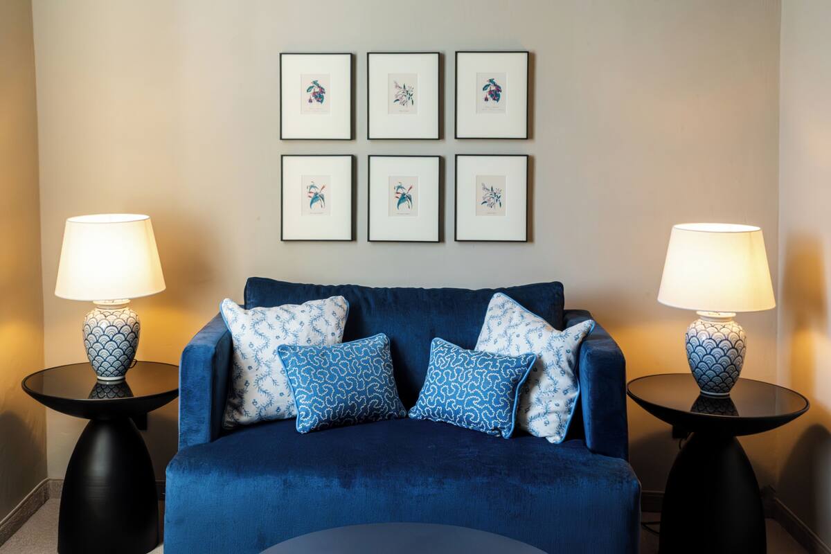 Un divano blu con cuscini decorati, due tavolini neri con lampade e sei opere d'arte incorniciate appese al muro.