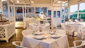 Un restaurante con mesas y sillas blancas, un chef preparando comida y vistas al exterior a través de amplios ventanales.