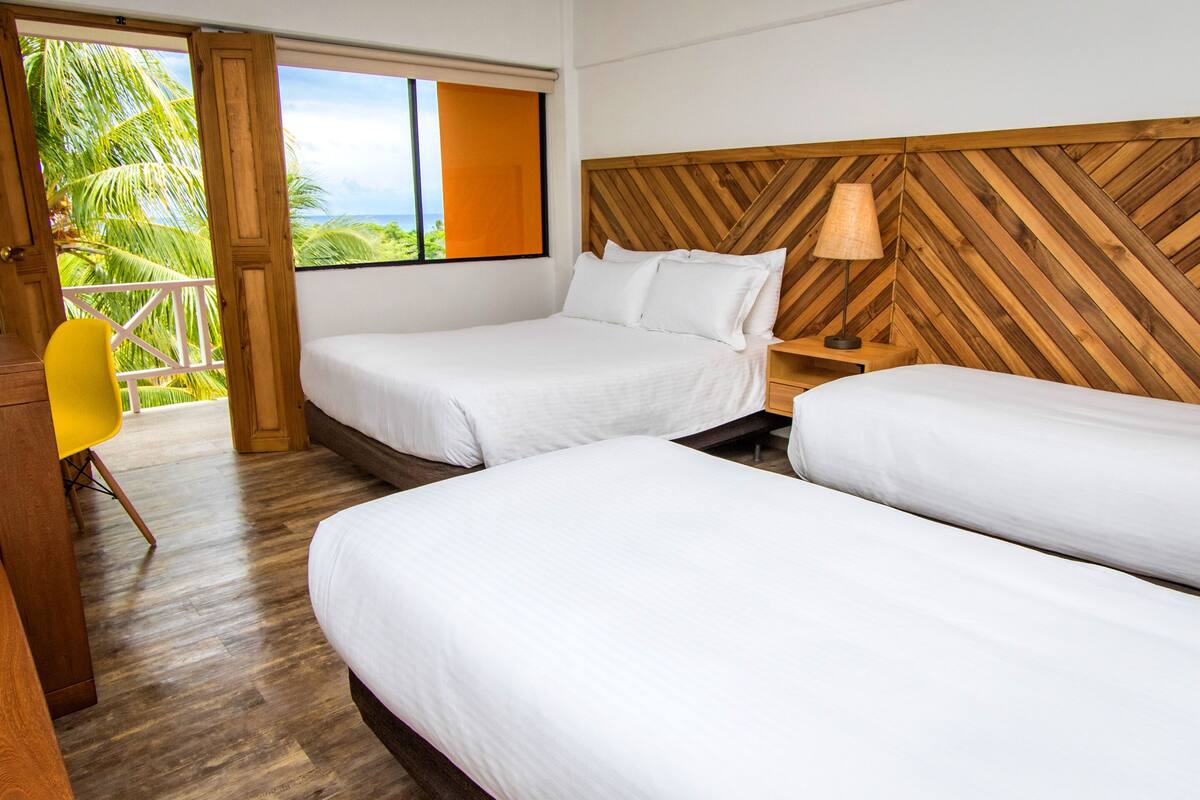 Ein Hotelzimmer mit zwei Betten, einem Holzkopfende, einem Nachttisch mit Lampe und Blick auf Palmen durch eine offene Tür.