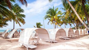 Una playa con tiendas de campaña blancas, sillas de descanso, palmeras y un cielo despejado.