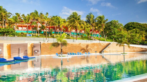 Una piscina con sillas reclinables azules, un paseo bordeado de palmeras y un edificio con balcones al fondo.