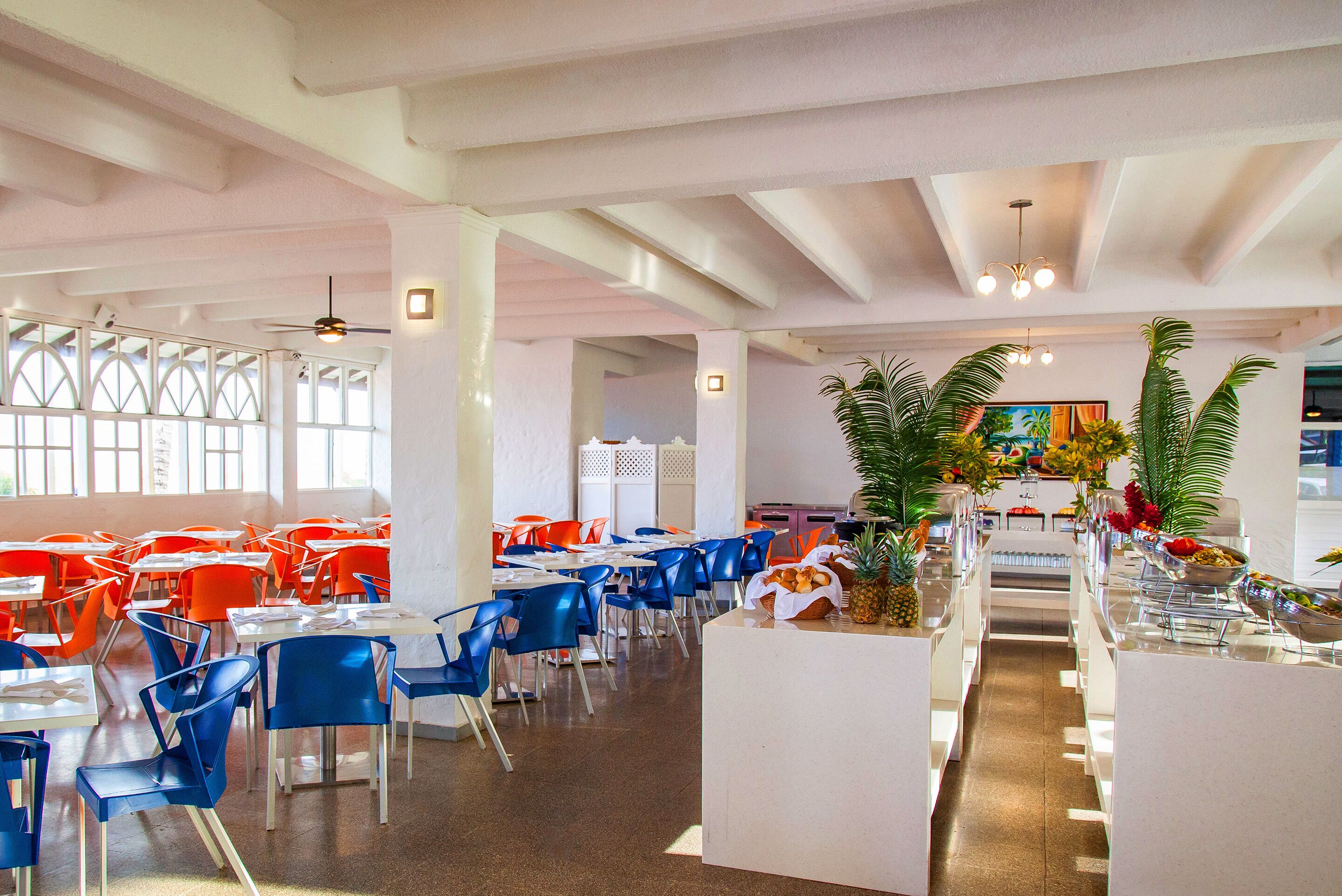 A spacious dining area with high ceilings and large windows, featuring blue and orange chairs and a central serving station with fresh fruits.