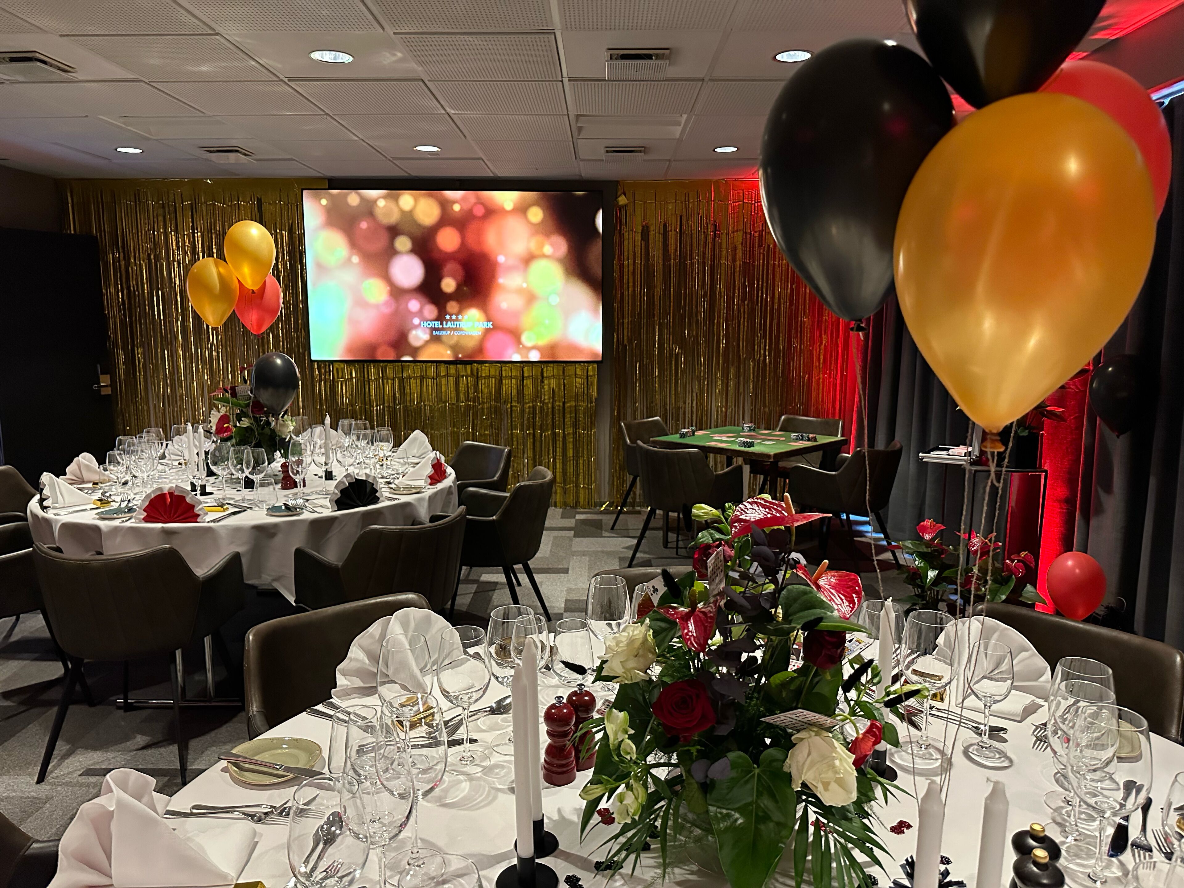 Un salón de banquetes con mesas redondas dispuestas para cenar, una pantalla grande al fondo y globos decorativos.