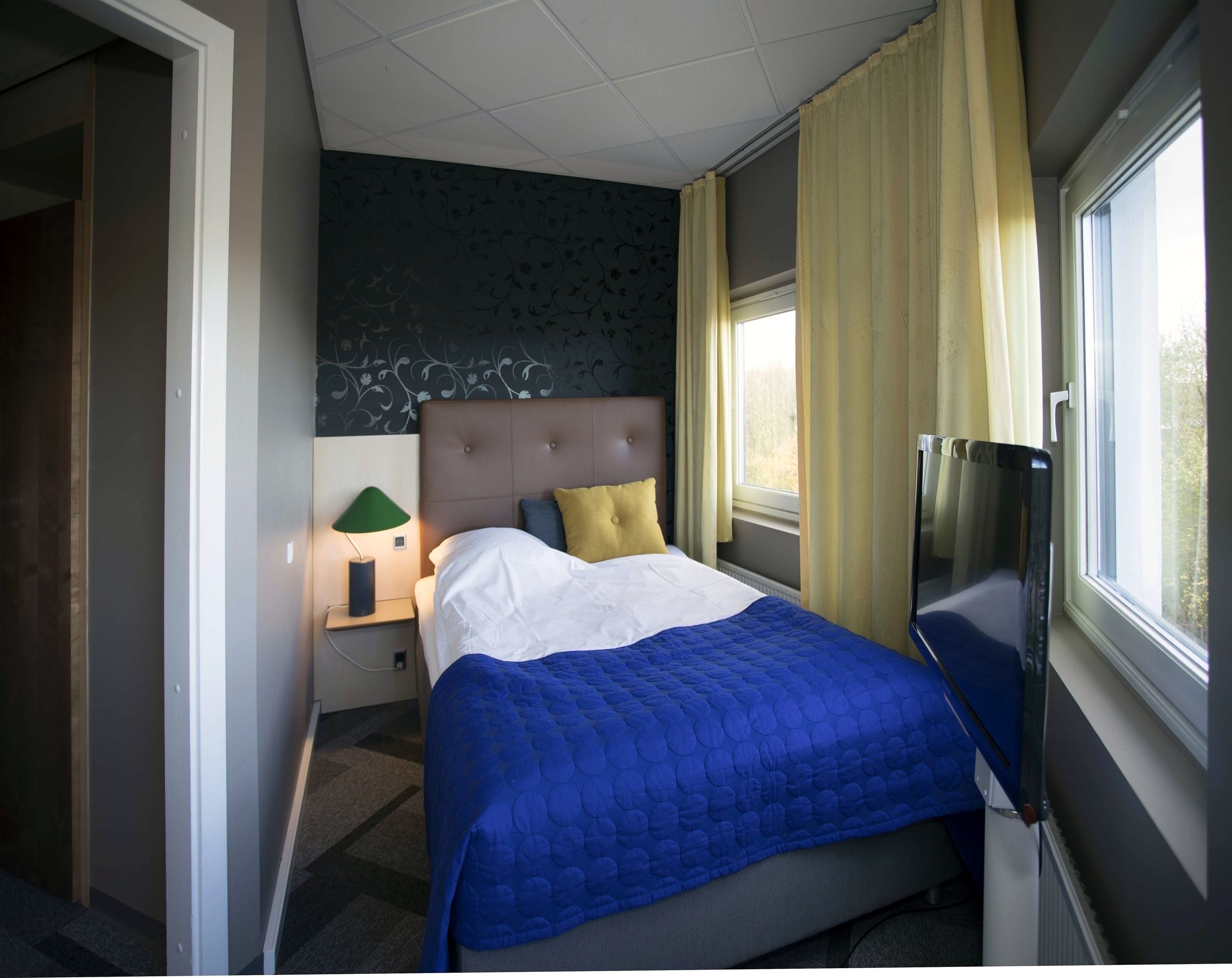Una habitación de hotel con una cama, televisión, mesita de noche, lámpara y ventana con cortinas.