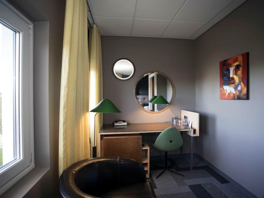 Ein Hotelzimmer mit Schreibtisch, zwei Stühlen, einem runden Spiegel und einem Gemälde an der Wand.