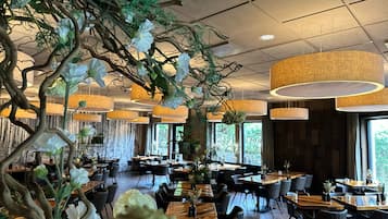 Un restaurante moderno con mesas y sillas de madera, lámparas colgantes y plantas de interior.