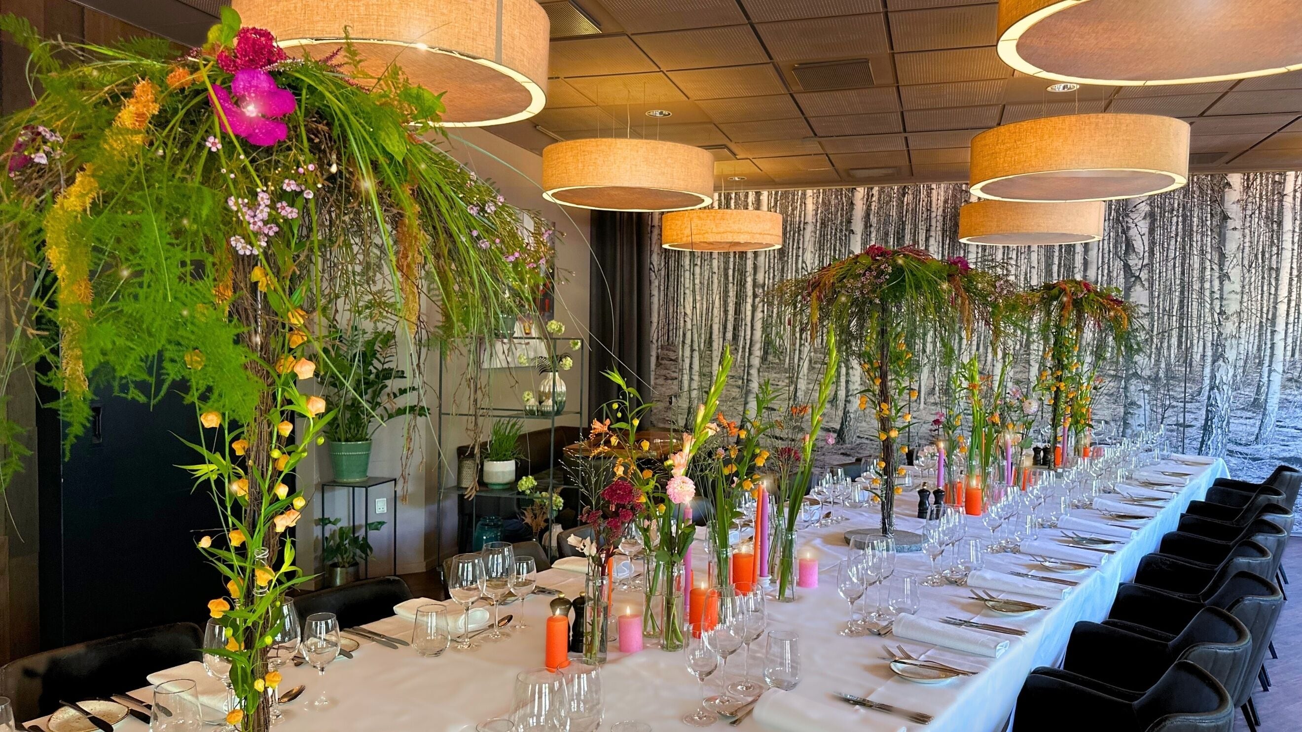 Ein langer Esstisch ist mit Gläsern und Besteck gedeckt, umgeben von hängenden Blumenarrangements und Pendelleuchten.