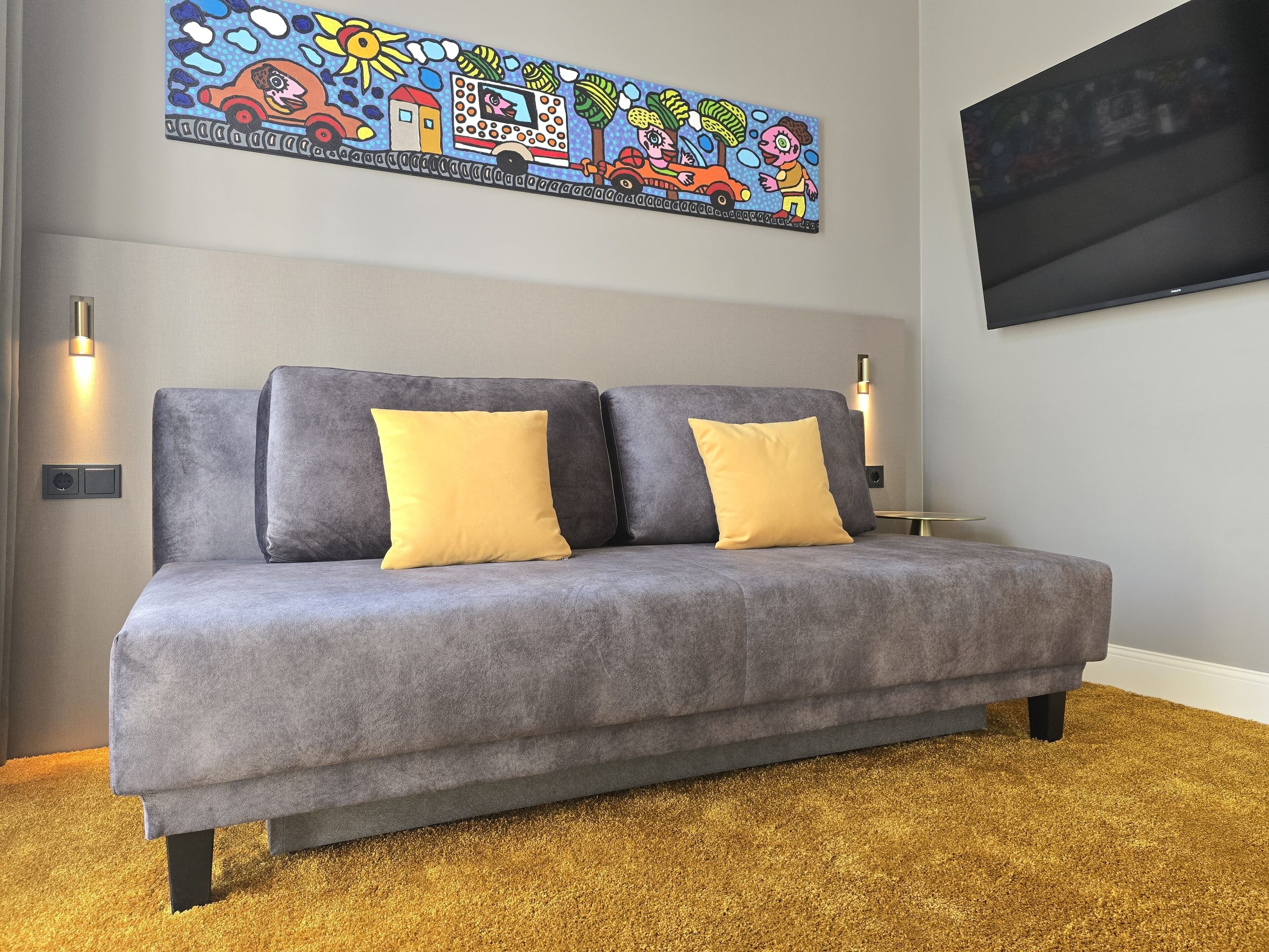 A grey sofa with two yellow cushions, a colorful abstract painting above, and a flat-screen TV on the wall.