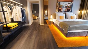 A modern hotel room with a large bed, a seating area with a sofa and a bench, a clothing rack, and a painting on the wall. - Lumière Boutiquehotel im Fürstenhof Leipzig (Leipzig)