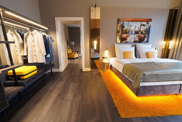 A modern hotel room with a large bed, a seating area with a sofa and a bench, a clothing rack, and a painting on the wall.