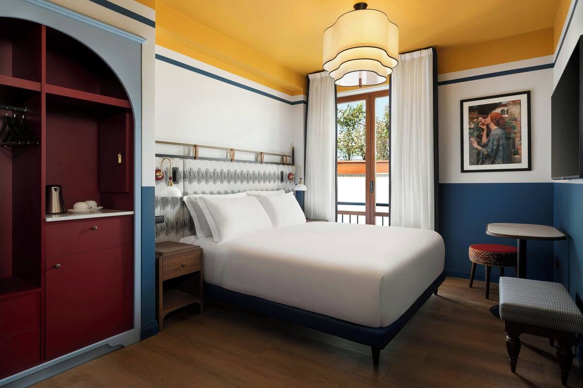 A hotel room with a bed, a bedside table, a wardrobe, a small round table, and a bench.