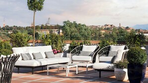 A rooftop terrace with outdoor seating, including a white sofa and wicker chairs, surrounded by potted plants and overlooking a cityscape.