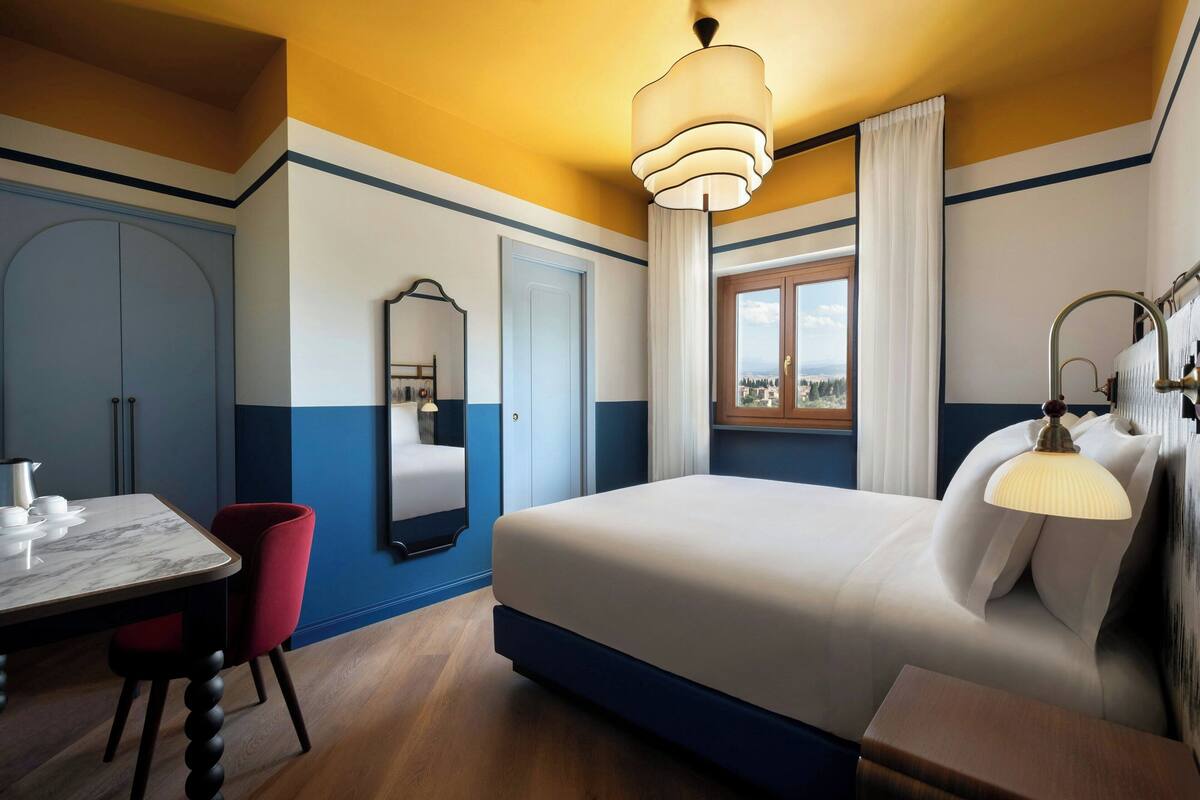 A hotel room with a bed, a desk with a chair, a large mirror, and a window with curtains.