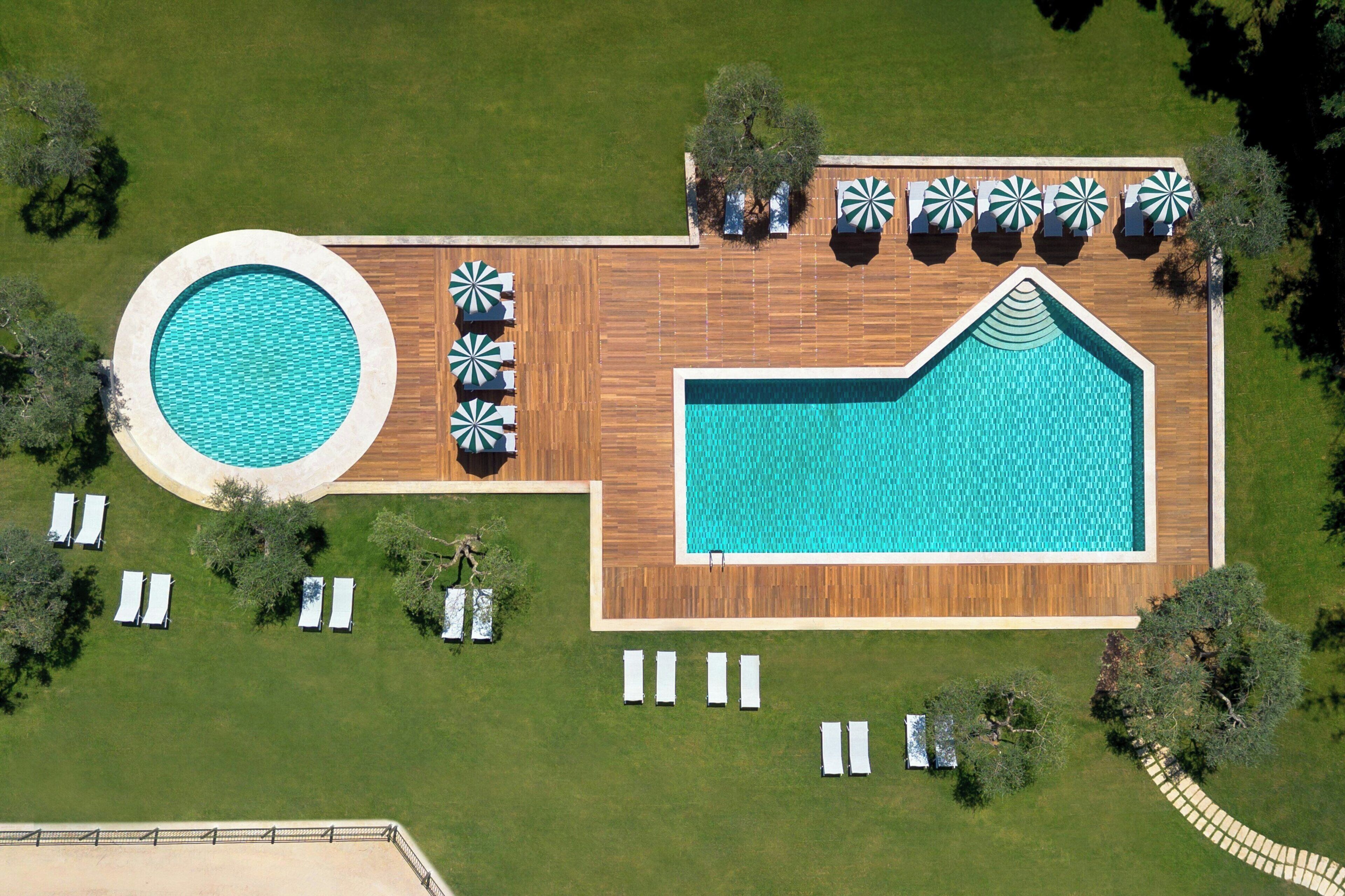 Vista aérea de um resort com uma piscina retangular grande, uma piscina circular e várias áreas de estar com guarda-sóis.