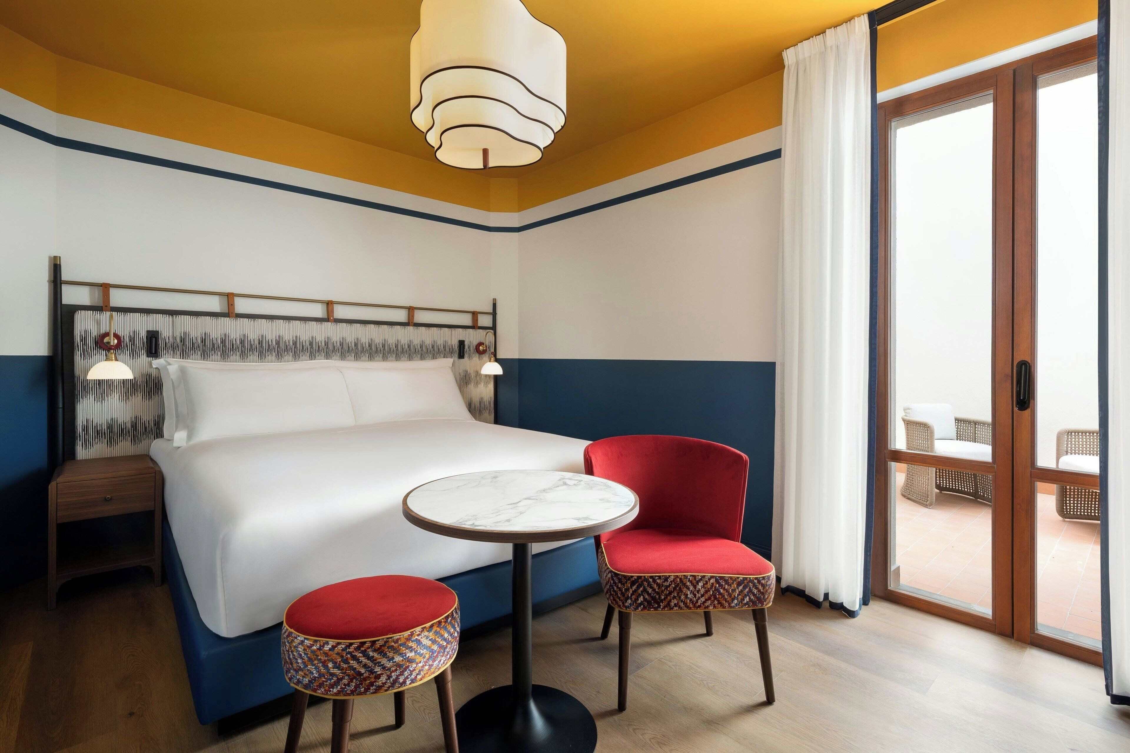 Hotellihuone, jossa on suuri sänky, pyöreä pöytä, kaksi punaista tuolia, pieni sivupöytä ja parveke, jossa on rottinkihuonekaluja.