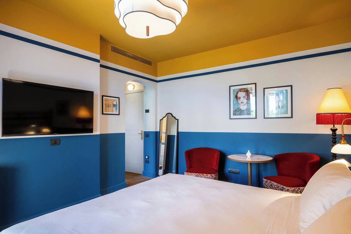 A hotel room with a bed, two red chairs, a small table, a TV, and artwork on the wall.