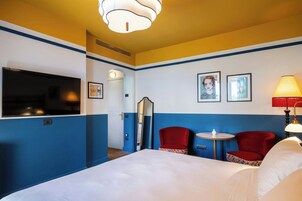 Una camera d'albergo con un letto, due poltrone rosse, un tavolino, una televisione e opere d'arte alle pareti.