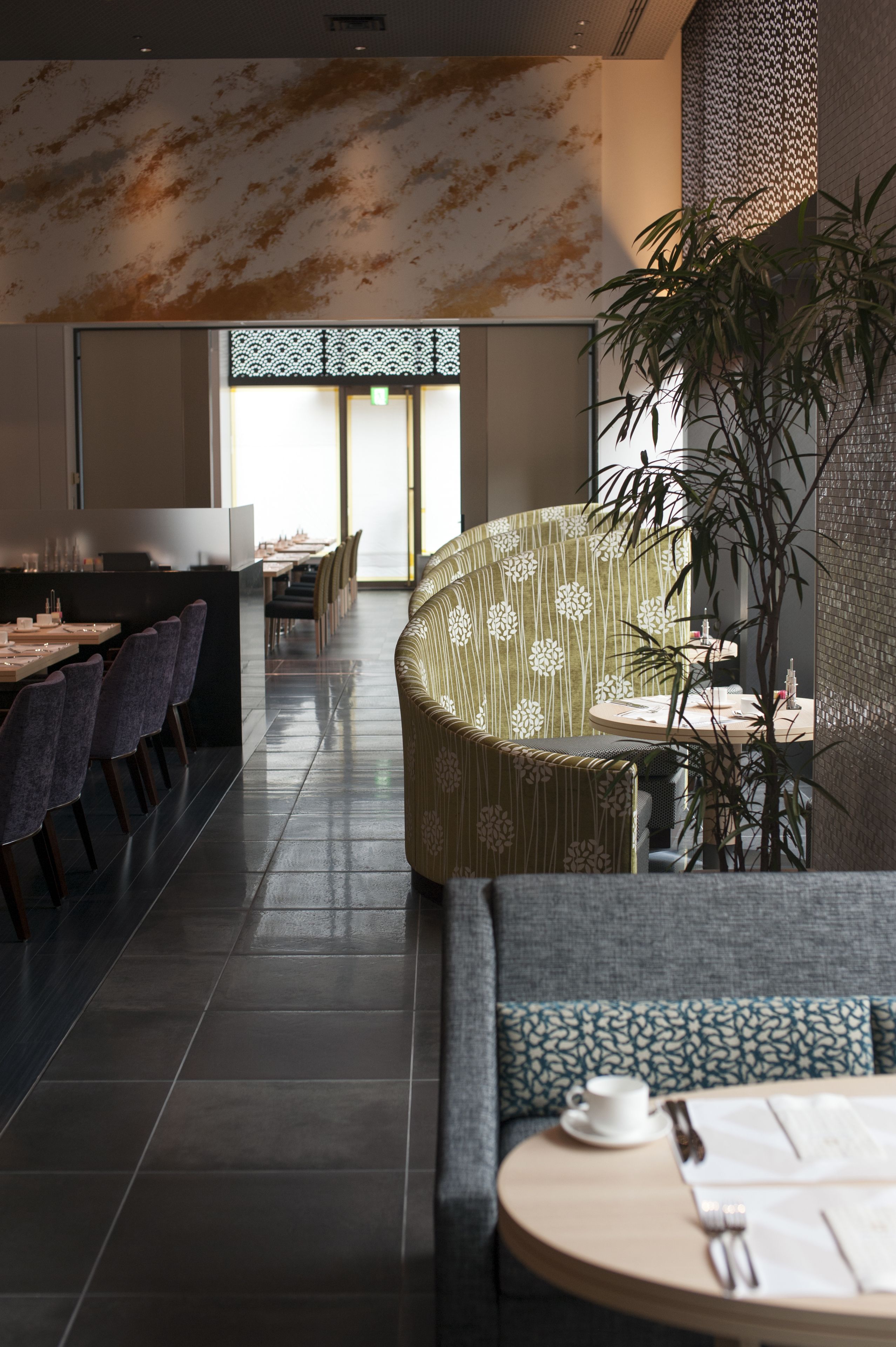 A modern dining area with a long table, chairs, and a sofa. There is a potted plant and a wall with a textured pattern.