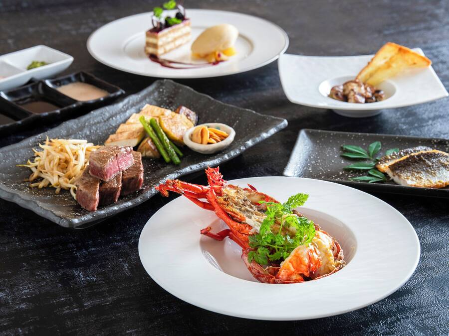 A variety of dishes on a dark table, including a lobster, steak, and other seafood.