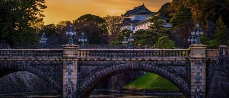 Un pont de pierre à deux arches enjambant une rivière paisible, menant à un bâtiment de style japonais traditionnel, doté d’un toit vert et de lanternes décoratives.