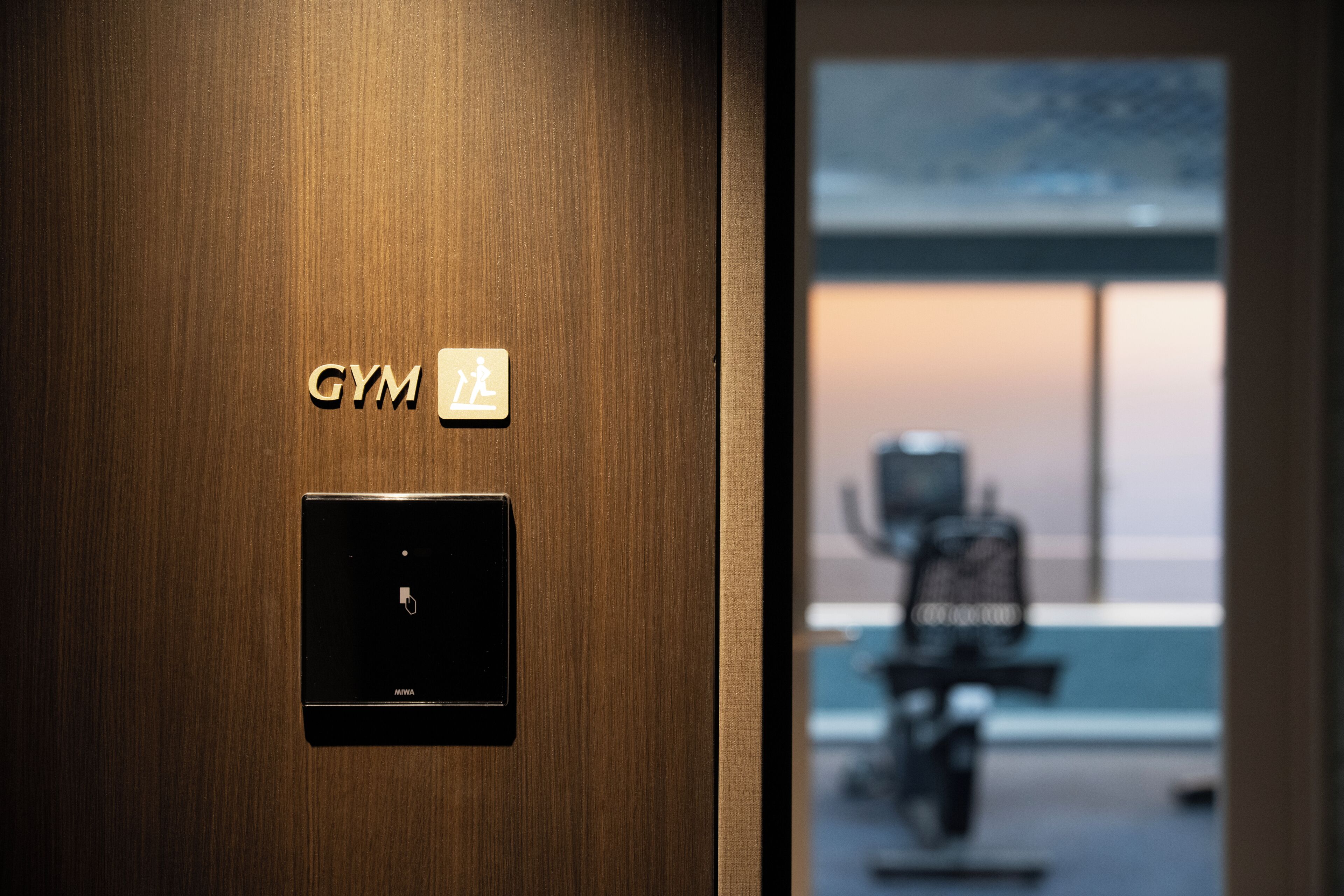 A wooden door with a 'GYM' sign and a control panel, leading to a room with a view of a building and a parked motorcycle.
