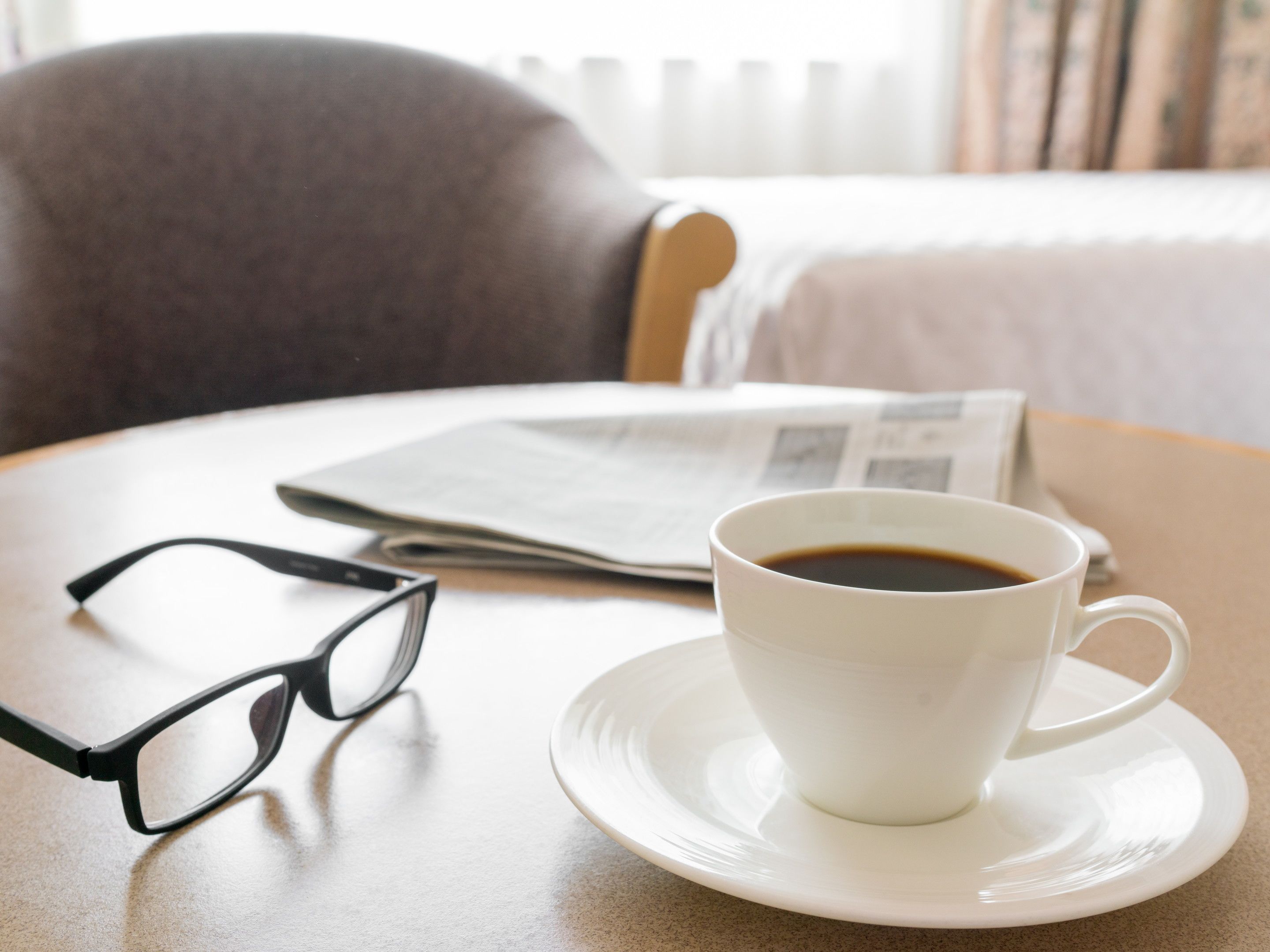A white cup of coffee on a saucer, a pair of glasses, and a newspaper on a round table.