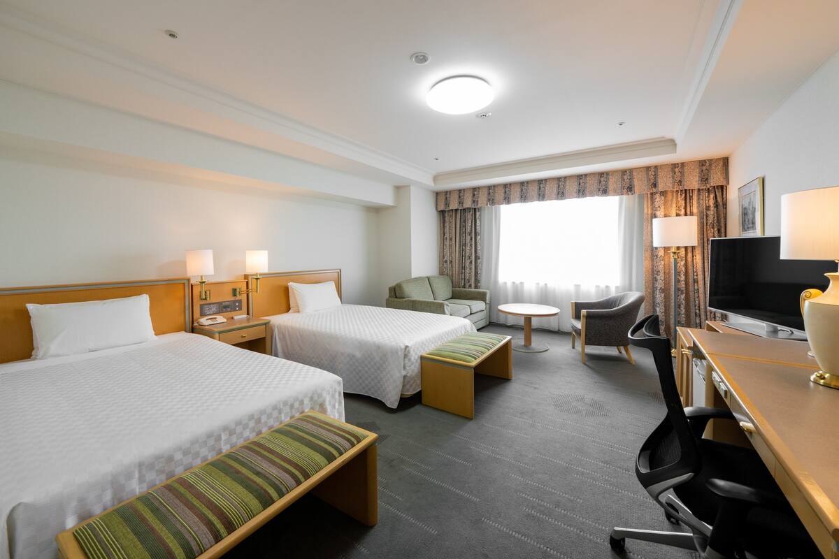 Une chambre d’hôtel avec deux lits, un bureau, une chaise, une télévision et une fenêtre avec des rideaux.