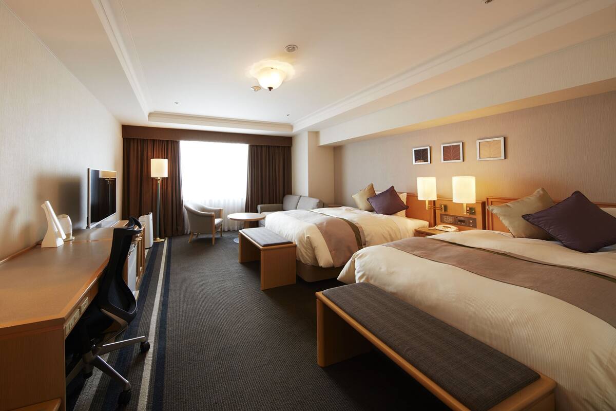 Une chambre d’hôtel avec deux lits, un bureau, une chaise, une télévision et une grande fenêtre avec des rideaux.