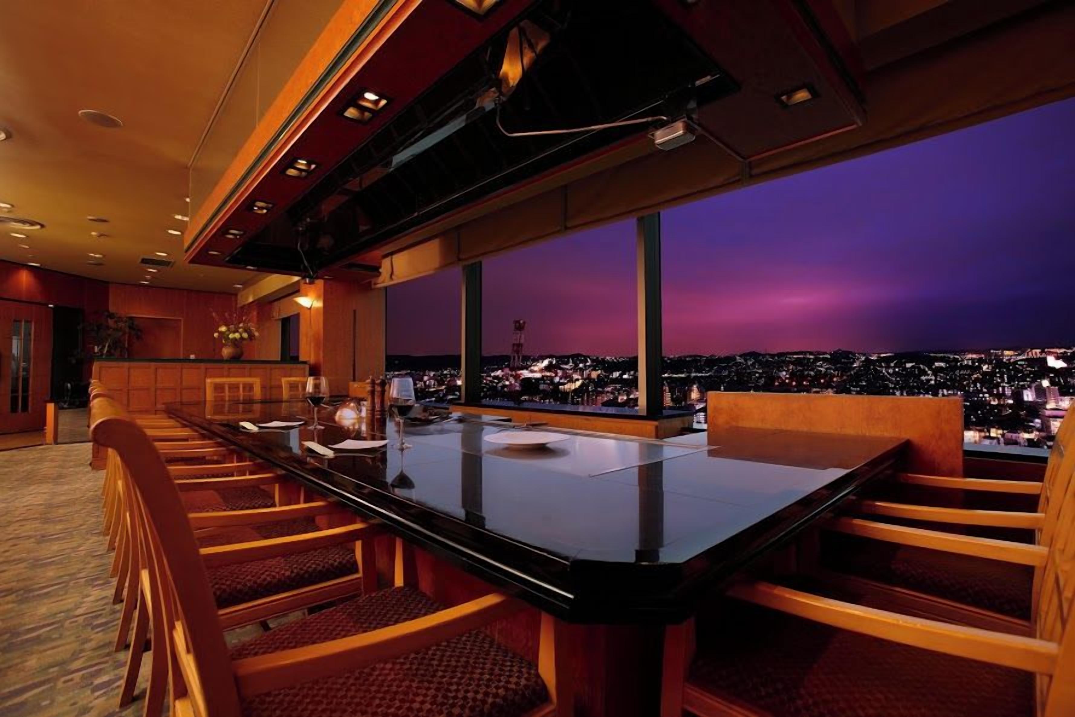 Un comedor con una mesa larga y sillas, ventanas amplias y vistas a la ciudad al anochecer.