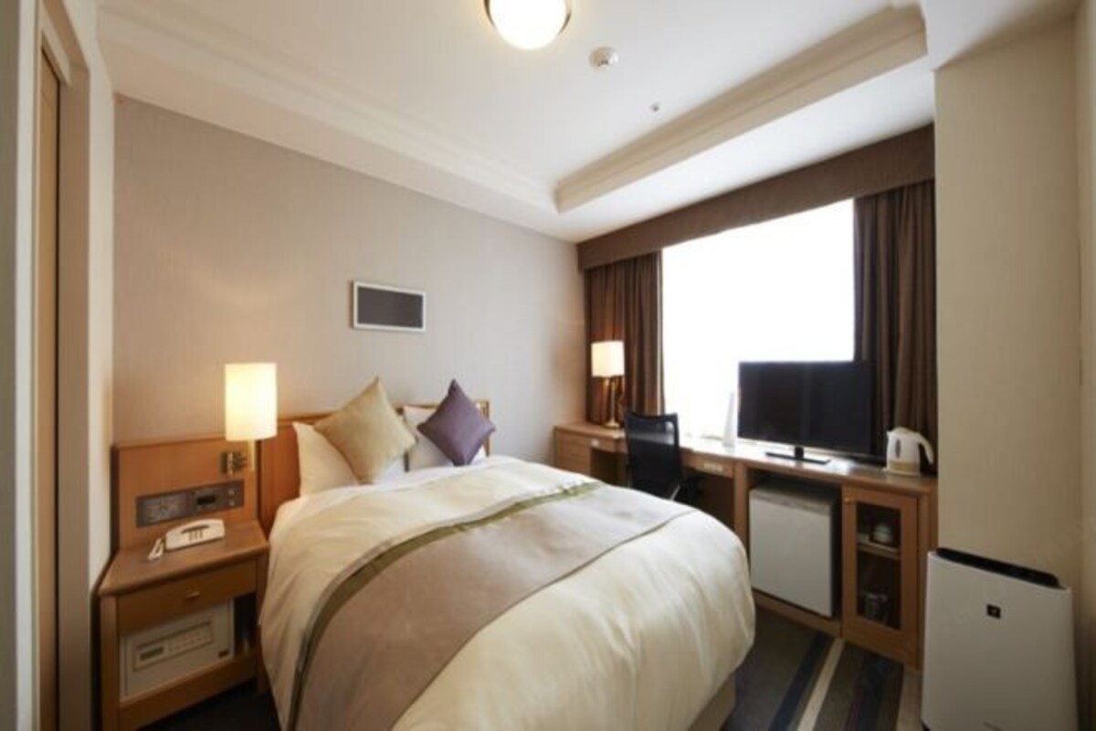 Una habitación de hotel con una cama grande, un escritorio con una computadora, un televisor y un ventanal con cortinas.