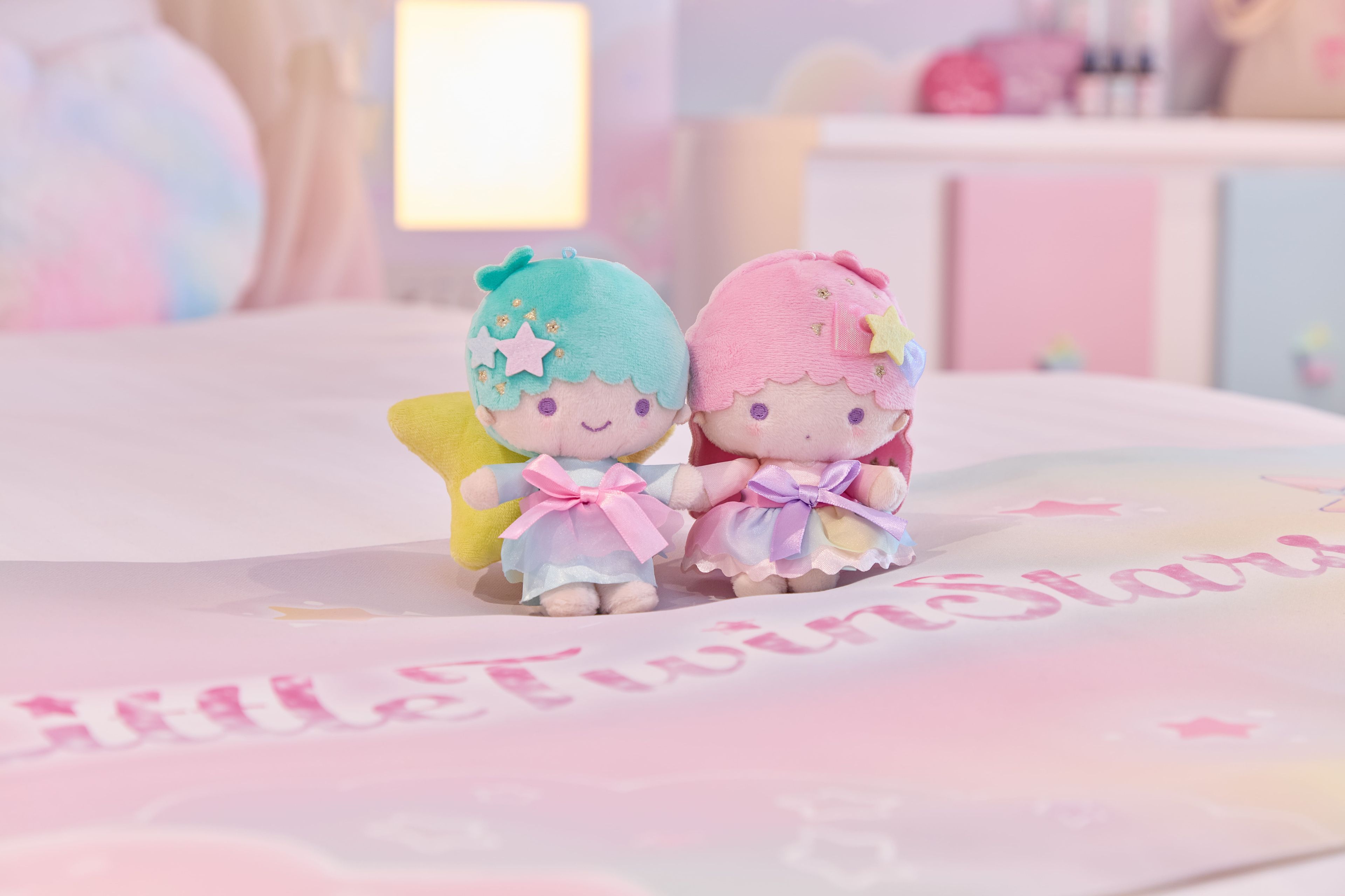 Two plush toys on a bed with a bedspread that has the text 'Little TwinStars'.