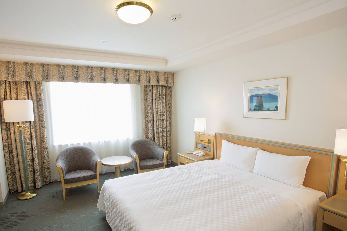 Une chambre d’hôtel comprenant un lit, deux fauteuils, une petite table, une table de chevet avec une lampe, un tableau au mur et une fenêtre avec des rideaux.